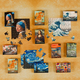A collection of Mini Masterpieces Jigsaw Puzzles by Professor Puzzle displayed on a textured surface. Various puzzle boxes feature famous artworks, including the Girl with a Pearl Earring, Starry Night, and The Wave. Some puzzle pieces are scattered and partially assembled, showcasing vibrant colors and intricate designs. Perfect for art lovers and puzzle enthusiasts.