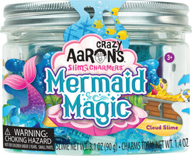 A clear jar filled with whimsical, blue cloud slime featuring colorful charms, including a mermaid tail and treasure chest, labeled 'Mermaid Magic' by Crazy Aaron's Slime Charmers. The jar has a warning label indicating a choking hazard for small parts.
