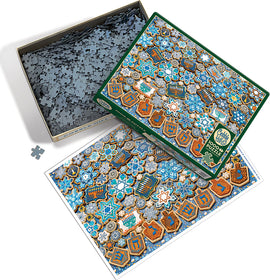 The image features the Cobble Hill Hanukkah Cookies jigsaw puzzle. The puzzle box is open and shows a colorful design with blue, gold, and white motifs resembling Hanukkah cookies, stars, dreidels, and menorahs. Scattered puzzle pieces are visible within the box, creating a festive holiday scene.