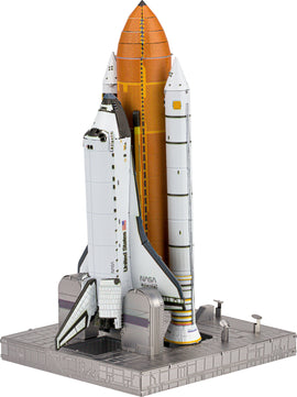 A detailed model kit of the Space Shuttle, with a white exterior and orange external tank. The model is displayed on a realistic launch pad base, showcasing the shuttle's wings, engines, and NASA markings.
