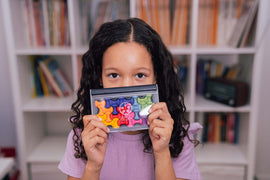 A child with curly hair smiling while holding the IQ Waves mechanical puzzle toy, which features colorful, interlocking pieces in a compact storage case. The background shows a cozy room with shelves filled with books.
