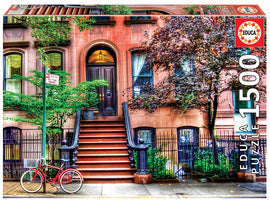 The GREENWICH VILLAGE jigsaw puzzle by Educa features a picturesque street scene with a classic brownstone building, decorative entrance, lush trees, and a vintage bicycle parked on the sidewalk. This 1500-piece puzzle captures the charm of New York City, perfect for puzzle enthusiasts and city lovers.