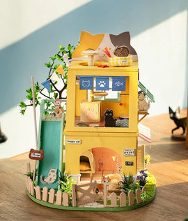 A detailed miniature cat house DIY kit by Robotime, featuring two levels with playful decor, a slide, trees, and cute cat figurines. The vibrant yellow structure has a welcoming sign and various cat-themed decorations, inviting imaginative play and creativity.