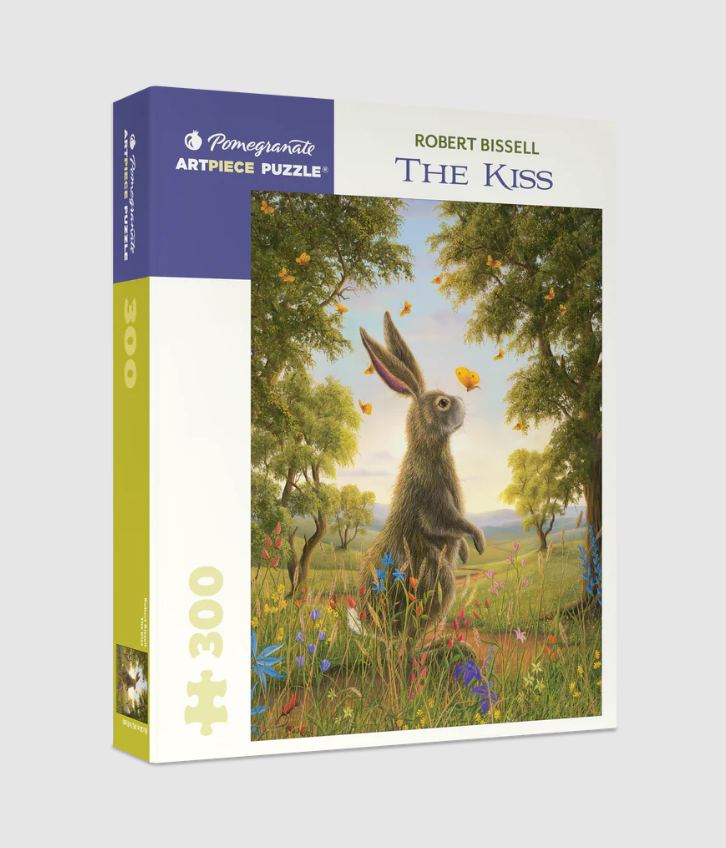 The Kiss jigsaw puzzle by Pomegranate shows a whimsical scene featuring a large, furry rabbit surrounded by colorful flowers and butterflies in a lush green landscape. The puzzle box displays the name 'The Kiss' by artist Robert Bissell, designed for 300 pieces.