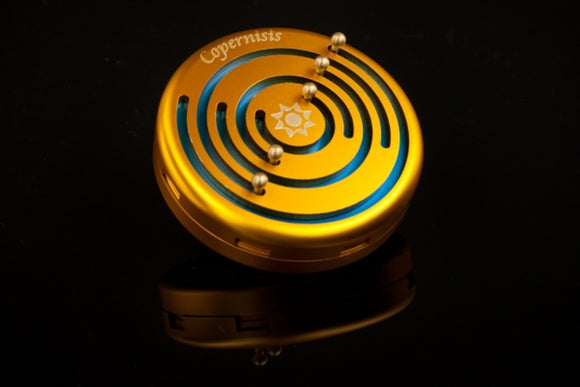 The Copernisis Gold & Blue mechanical puzzle by Sonicwarp features a stunning circular design with a vibrant gold finish. It has concentric grooves and four small metal balls placed strategically, inviting users to engage in a hands-on challenge while also enhancing fine motor skills and cognitive abilities. The reflective surface beneath adds a touch of elegance.