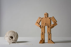 The Cubebot Micro - Julien, a charming mechanical puzzle, is displayed standing upright. It features an innovative wooden design with articulated limbs, allowing for various poses. Next to it is a ball of beige string, creating a simple and earthy aesthetic. Ideal for both kids and adults, this puzzle adds a playful touch to any space.