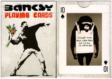 A deck of playing cards featuring Banksy artwork. The front displays a figure throwing flowers, while the reverse shows a monkey holding a sign that reads 'Laugh now, but one day we'll be in charge.' The deck is designed by Piatnick and showcases urban art themes.