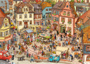 A vibrant jigsaw puzzle illustration titled 'Market Place' by Göbel & Knorr, featuring a bustling town square filled with people, market stalls, colorful buildings, and various activities such as dancing, shopping, and street performances. Lush greenery and detailed architectural elements enhance the lively atmosphere of the scene.
