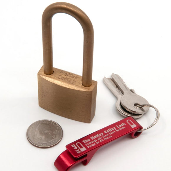 The HoKey CoKey Lock by Two Brass Monkeys, featuring a brass mechanical padlock with a U-shaped shackle. Beside it are two keys on a red key holder with 'The HoKey CoKey Lock' inscribed, along with a quarter for size reference.