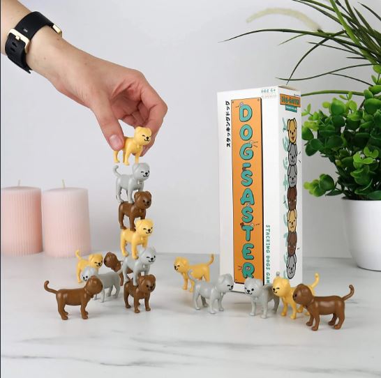 A hand stacking small, colorful dog figurines in a playful game called Dogsaster by Gift Republic. The box displays vibrant graphics and a fun title, alongside decorative candles and a green plant.