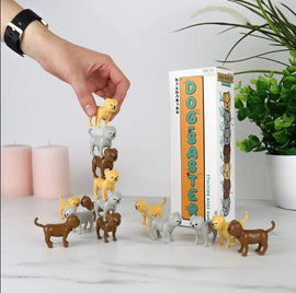 A hand stacking small, colorful dog figurines in a playful game called Dogsaster by Gift Republic. The box displays vibrant graphics and a fun title, alongside decorative candles and a green plant.