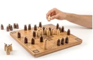 A hand moves a light-colored piece on a Hnefatafl game board. The board features wooden squares and intricately designed pieces in two colors, representing a strategic two-player game.