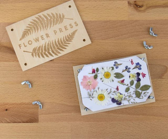 A wooden flower press set from Gift Republic, featuring a beautifully engraved 'FLOWER PRESS' label. The kit includes a wooden frame, pressed flowers, and securing screws displayed on a wooden surface, ideal for DIY floral crafts.