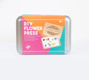 A DIY Flower Press kit packaged in a colorful tin box. The front features the words 'DIY Flower Press' in bold white letters against an orange background, with images of pressed flowers and a wooden board labeled 'Flower Press'. Suitable for ages 8 and up, this kit allows users to create beautiful pressed flower designs for decoration.