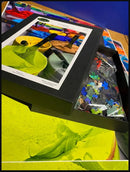 A Custom Jigsaw puzzle from Eureka Puzzles displayed in its box. The front of the box features vibrant paint colors, while an assortment of colorful jigsaw pieces is visible in the bag beside it. The background shows an artistic layout with more colorful textures.
