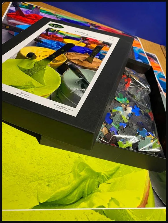 A Custom Jigsaw puzzle from Eureka Puzzles displayed in its box. The front of the box features vibrant paint colors, while an assortment of colorful jigsaw pieces is visible in the bag beside it. The background shows an artistic layout with more colorful textures.