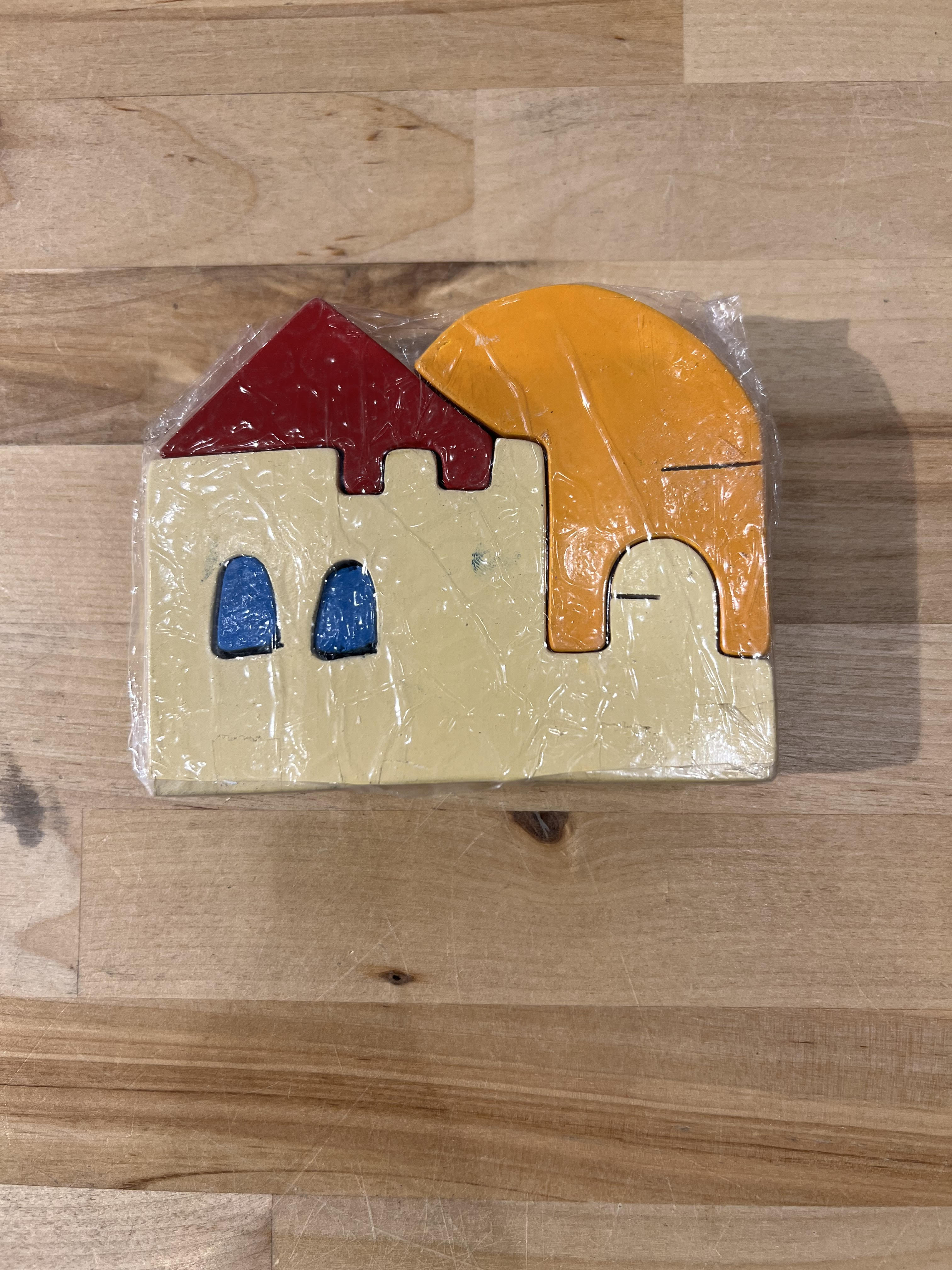 A colorful wooden jigsaw puzzle featuring a stylized house with a red roof, blue windows, and a large orange sun. The pieces are made from natural wood and are intricately shaped, showcasing vibrant colors. Wrapped in clear plastic, the puzzle emphasizes creativity and tactile play for children.