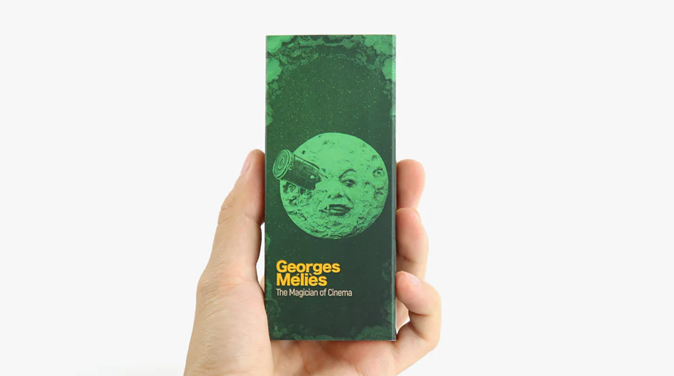 A person holding a flipbook titled 'Georges Melies: The Magician of Cinema' by Boku Flipbooks. The cover features a green-tinted illustration of a moon with a face, reminiscent of Melies' classic cinematic style. The book's title is printed in bold yellow letters, highlighting its connection to the influential filmmaker.