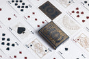 A close-up view of Bicycle Cypher Cards displayed on a surface. The deck features intricate designs in gold and black, showcasing various playing cards laid out around the box. The cards include a mix of hearts and spades, with notable face cards visible. This high-quality deck designed by US Playing Card Co. combines stunning artwork with gameplay functionality.