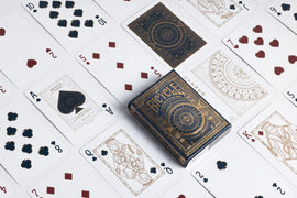 A close-up view of Bicycle Cypher Cards displayed on a surface. The deck features intricate designs in gold and black, showcasing various playing cards laid out around the box. The cards include a mix of hearts and spades, with notable face cards visible. This high-quality deck designed by US Playing Card Co. combines stunning artwork with gameplay functionality.