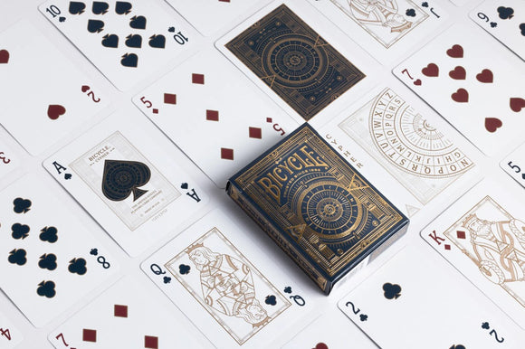 A close-up view of Bicycle Cypher Cards displayed on a surface. The deck features intricate designs in gold and black, showcasing various playing cards laid out around the box. The cards include a mix of hearts and spades, with notable face cards visible. This high-quality deck designed by US Playing Card Co. combines stunning artwork with gameplay functionality.
