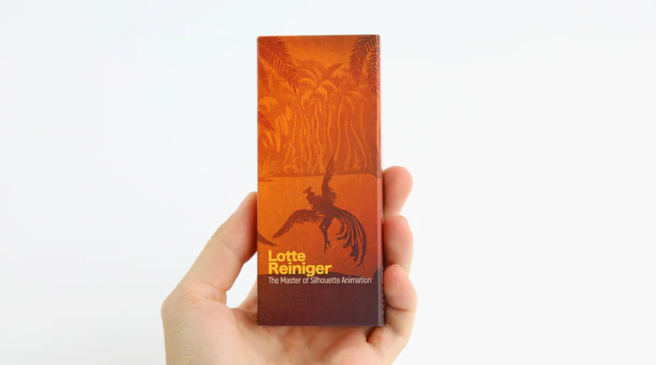 A hand holding a FlipBoku product featuring Lotte Reiniger, known as the Master of Silhouette Animation. The box is designed with warm orange and earthy tones, illustrating silhouette designs reminiscent of Reiniger's animation style, set against a backdrop of palm trees.