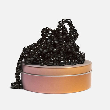 A collection of black, smooth, bean-shaped pieces playfully tangled on top of a round, pastel-colored tin. The tin has a metallic sheen and is slightly wider at the base, giving it a modern aesthetic. This visual captures the playful essence of the SPEKS Crags: Dandy product, inviting curiosity and engagement.