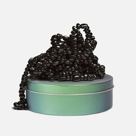 The product 'Crags: Tranquility' by SPEKS is a unique fidget item consisting of many black, smooth, bead-like pieces loosely gathered on top of a round, gradient-colored tin with an iridescent finish. The tin has a subtle transition from green to blue, enhancing the overall aesthetic of the product while showcasing its playful and stress-relieving nature.
