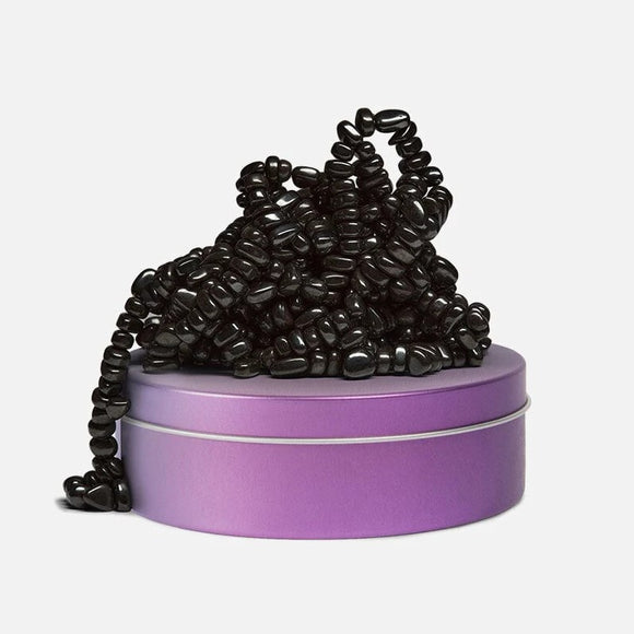A playful assortment of black, bean-shaped pieces stacked in a casual pile, partially resting on a purple tin container. The pieces are smooth and shiny, creating a sensory and tactile experience, ideal for fidgeting or creative exploration. The purple tin adds a pop of color and a convenient storage option.