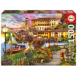 A beautifully illustrated jigsaw puzzle titled 'Italian Promenade' by Educa, featuring a picturesque lakeside scene with colorful houses, vibrant flowers, a classic boat, and a serene sunset. This 1500-piece puzzle captures the charm of Italian architecture and nature, inviting puzzlers to explore its intricate details.