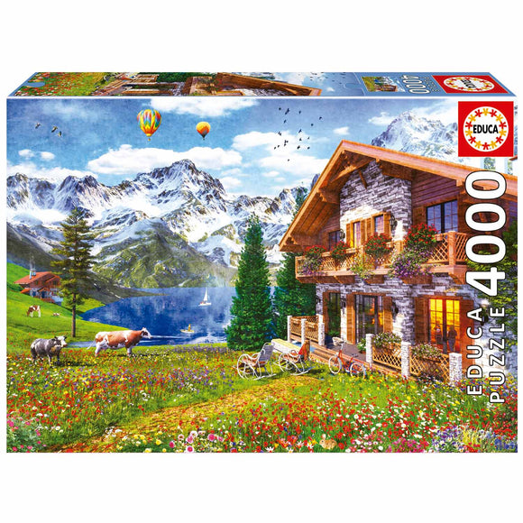 A beautifully illustrated jigsaw puzzle titled 'Chalet in the Alps' by Educa. The scene features a charming chalet surrounded by vibrant wildflowers, green meadows, and majestic snow-capped mountains. Hot air balloons float in the sky above. A serene lake reflects the landscape, with cows grazing in the foreground and a small sailboat on the water. This detailed artwork captures the tranquility and beauty of the Alpine region.