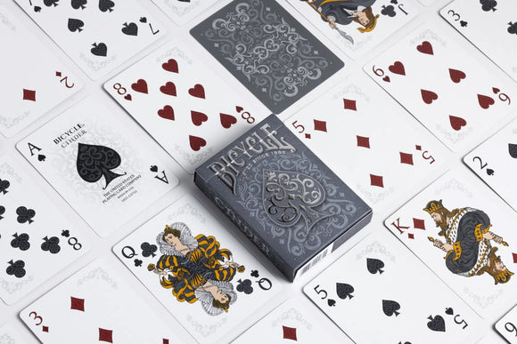 Bicycle Cinder playing cards by US Playing Card Co. The box is prominently displayed in a dark grey color with intricate designs. Surrounding the box are several opened playing cards showing various suits, including hearts, spades, and diamonds, featuring artistic illustrations.