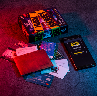 The Evidence Hush Money Game by Professor Puzzle displayed on a textured surface. The packaging features bold, colorful graphics. Nearby, a red wallet holds various cards, and a smartphone shows a game screen. The setting has a dramatic lighting effect with blue and pink hues.