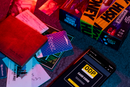 Image of the Evidence Hush Money Game by Professor Puzzle. The colorful box is displayed alongside various cards, business documents, and a smartphone showing a case code. The game is set on a textured surface with a mix of detective elements suggesting intrigue and mystery.