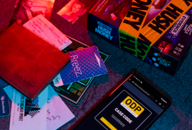 Image of the Evidence Hush Money Game by Professor Puzzle. The colorful box is displayed alongside various cards, business documents, and a smartphone showing a case code. The game is set on a textured surface with a mix of detective elements suggesting intrigue and mystery.
