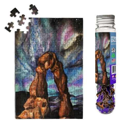 A finished 150-piece jigsaw puzzle depicting Arches National Park with a colorful night sky. The puzzle is shown alongside its packaging, a clear bottle with a decorative label. Several puzzle pieces are scattered around the image.