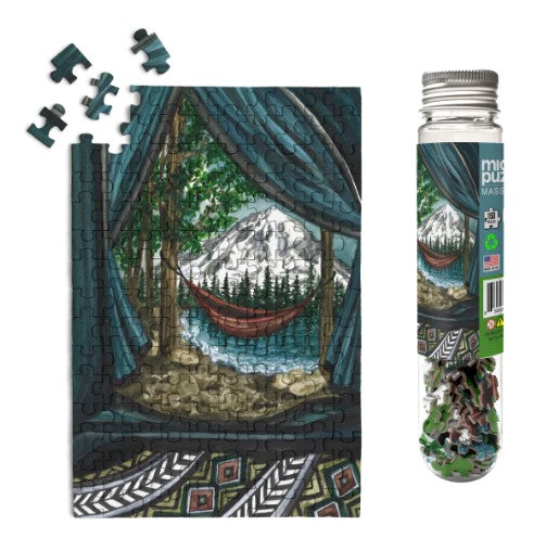 A close-up of the Mt. Rainier National Park jigsaw puzzle by Micro Puzzle. The image features a scenic view of a mountainside with a hammock, framed by trees and a cozy cabin interior. The puzzle pieces are arranged both on the puzzle and nearby, with the packaging visible in a glass tube.