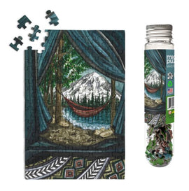 A close-up of the Mt. Rainier National Park jigsaw puzzle by Micro Puzzle. The image features a scenic view of a mountainside with a hammock, framed by trees and a cozy cabin interior. The puzzle pieces are arranged both on the puzzle and nearby, with the packaging visible in a glass tube.