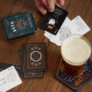 A stylish set of Beer Playing Cards by Gentlemen's Hardware displayed on a wooden table. The tin case features a hop design and the word 'BEER.' Nearby, a hand is shown holding a black playing card, while several other cards and a glass of beer sit on the table, enhancing the game's social atmosphere.