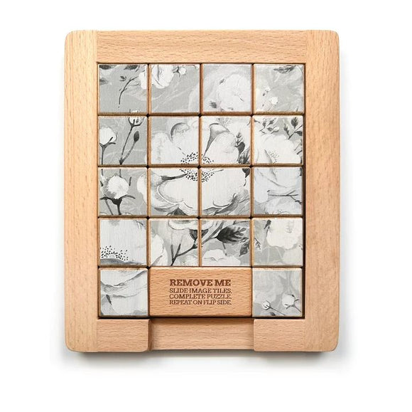 A wooden mechanical sliding puzzle featuring black and white floral designs. The puzzle consists of a grid of tiles that can be rearranged to complete the image. The base has a wood finish with an instruction label that says 'REMOVE ME' for interaction, highlighting its playful nature. The background is minimalist to emphasize the puzzle's intricate details.