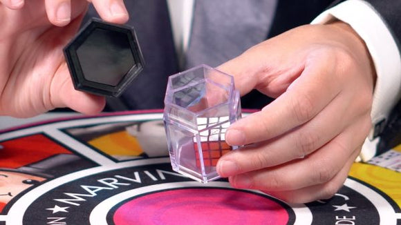 A close-up of a magician's hand holding a transparent hexagonal container and a black lid. The background features a colorful magic-themed board. The setup suggests a performance involving a Rubik's Cube magic set by Marvin's Magic.