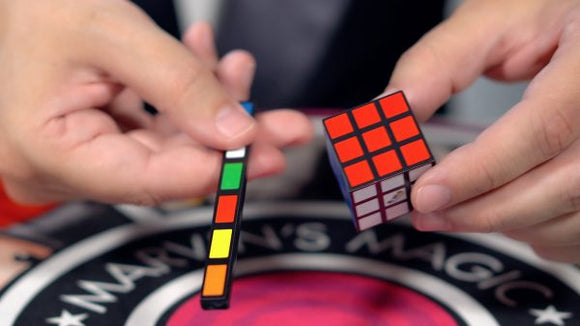 A person holding a Rubik's Cube and a mini version of a color-coded puzzle piece, demonstrating the Rubik's Cube Magic Set by Marvin's Magic. The vibrant colors of the cube are prominently displayed against a circular Marvin's Magic branded mat.