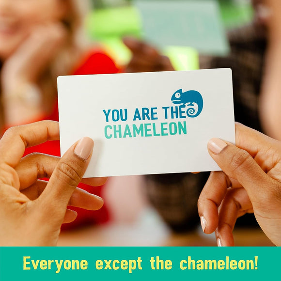A close-up of a hand holding a card that reads 'YOU ARE THE CHAMELEON' with a chameleon logo. The background shows blurred figures of people in a lively game setting, indicating a social atmosphere.