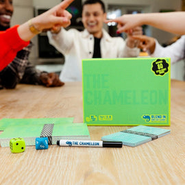 The Chameleon board game by Big Potato Games displayed on a wooden table. The game includes green and blue gameplay components, two colorful dice, a black marker, and a box featuring a vibrant design. People can be seen in the background, engaging in the game with playful gestures.