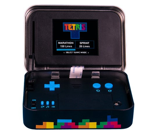 Tetris Arcade In A Tin by Fizz Creations Inc. opens to reveal a compact gaming device with a small screen displaying the Tetris logo and game mode options. The device features blue buttons for gameplay and a colorful Tetris block design on the bottom. Perfect for nostalgic gaming on the go.