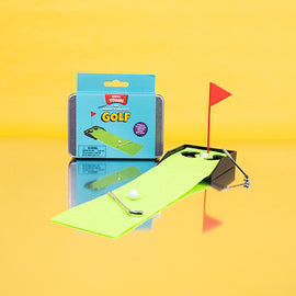 Teeny Town Golf game by Fizz Creations Inc. features a mini golf setup with a green felt putting surface, a small golf ball, a club, and a red flag. The packaging background is light blue, displaying instructions and brand details, set against a bright yellow backdrop.