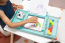 A child playing with the NugLug Complete Set. The product features a teal case open on a table, displaying colorful geometric tiles and letter pieces. The child is focused on assembling a design on a card labeled 'Difficulty 1 star' while behind them is a drawing marked 'HUMAN'. The table has a patterned rug underneath.