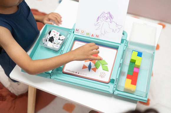A child playing with the NugLug Complete Set. The product features a teal case open on a table, displaying colorful geometric tiles and letter pieces. The child is focused on assembling a design on a card labeled 'Difficulty 1 star' while behind them is a drawing marked 'HUMAN'. The table has a patterned rug underneath.