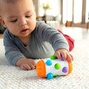 A baby in a gray onesie is playing on a soft rug, reaching for a colorful toy named Rolio by Fat Brain. The toy features an array of colorful buttons and shapes, designed to stimulate sensory play and motor skills.