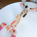 Two players engaging in a fun game of VolleyShot, a wooden game designed by Fat Brain. Each player holds a launcher, aiming to shoot a small ball through the center hole on a long, narrow wooden board. The game features colorful orange and black elements, and is set up on a white table, highlighting its interactive and competitive nature.